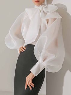 a woman is wearing a white blouse with an oversized bow and puff sleeves. the blouse is made of sheer fabric, and the sleeves are very voluminous. the woman is wearing black pants and is standing in front of a white background. she looks stylish and elegant.