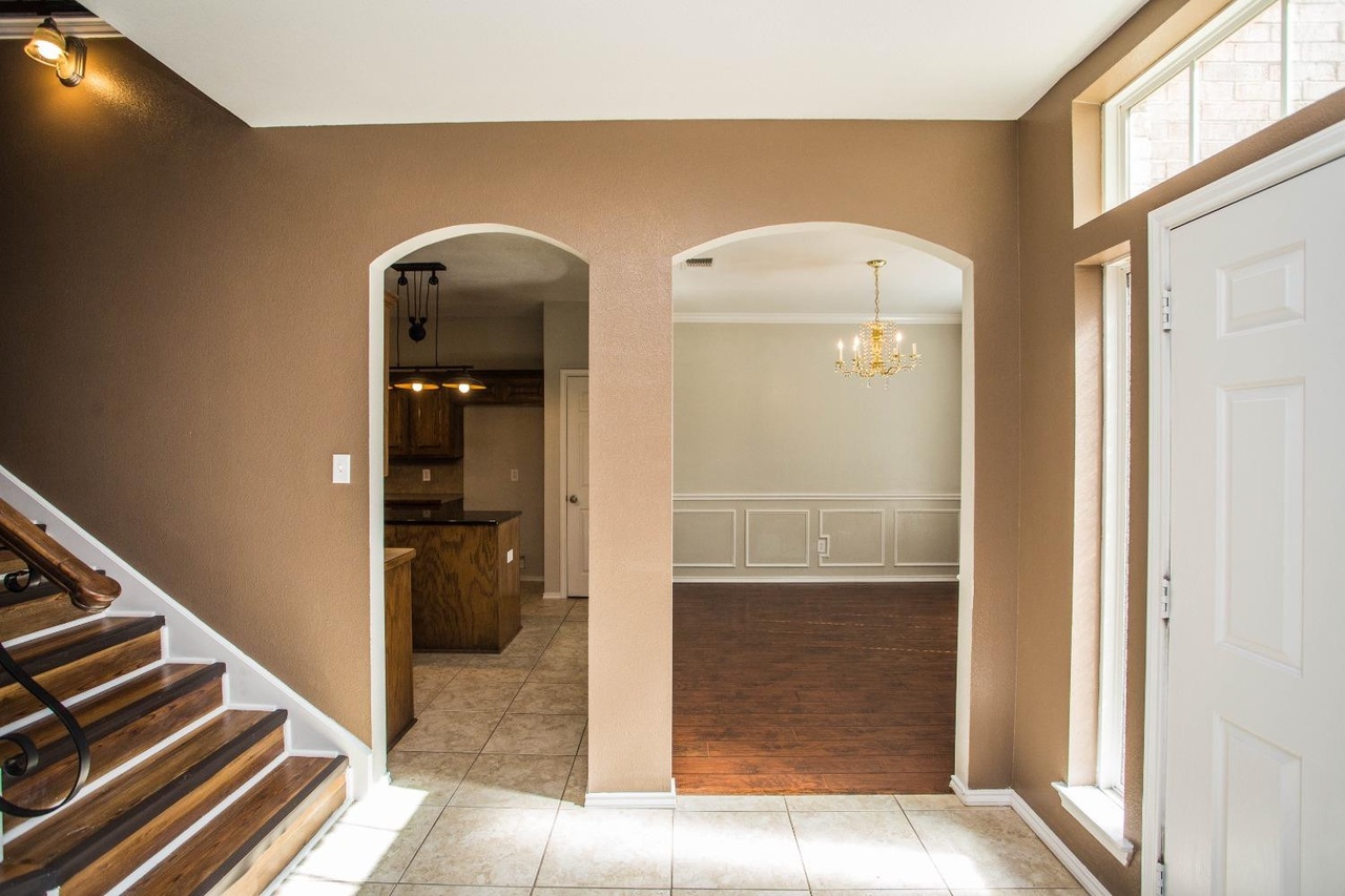interior view of an entryway with archways leading to a kitchen and dining room. a staircase is to the left and a white door and window are to the right. the walls are beige and the floors are tile in the entryway and kitchen and hardwood in the dining room. a chandelier hangs in the dining room.