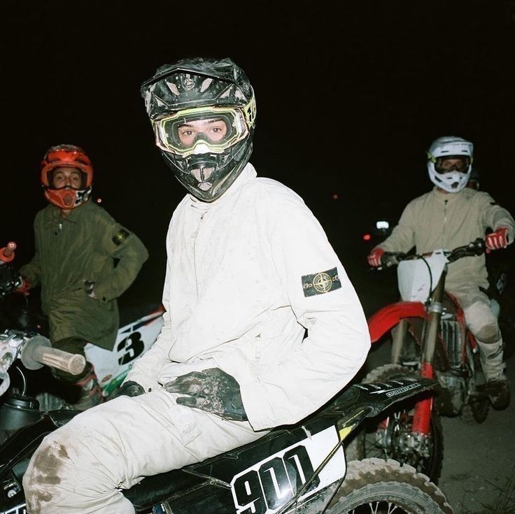 three young men in motocross gear are riding their motorcycles at night. the man in the center is wearing a white jacket with a black logo and a black helmet with goggles. he is in focus and looking at the camera. the man on the left is wearing a green jacket and red helmet, and he is out of focus. the man on the right is wearing a brown jacket and a white helmet, and he is also out of focus. they are all riding on dirt bikes and the scene is set at night. the ground is dirt, and the motorcycles are kicking up dirt as they ride.