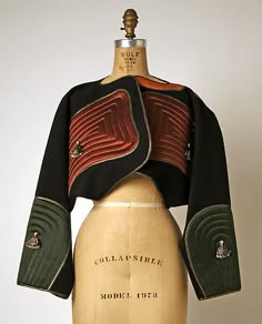 a vintage, 1970sstyle jacket on a dress form. the jacket is black with red, green and gold accents. the jacket is cropped, with long sleeves. the sleeves are finished with a green and gold trim, and the jacket features a collarless, highnecked design. the jacket is made of a thick, textured fabric. the jacket is designed to fit close to the body, and it is cinched in at the waist. the dress form is made of a white, smooth material. the dress form is also standing on a white base. the dress form is positioned in front of a white background. the dress form is an accurate representation of the human body, and it is used to display clothing.