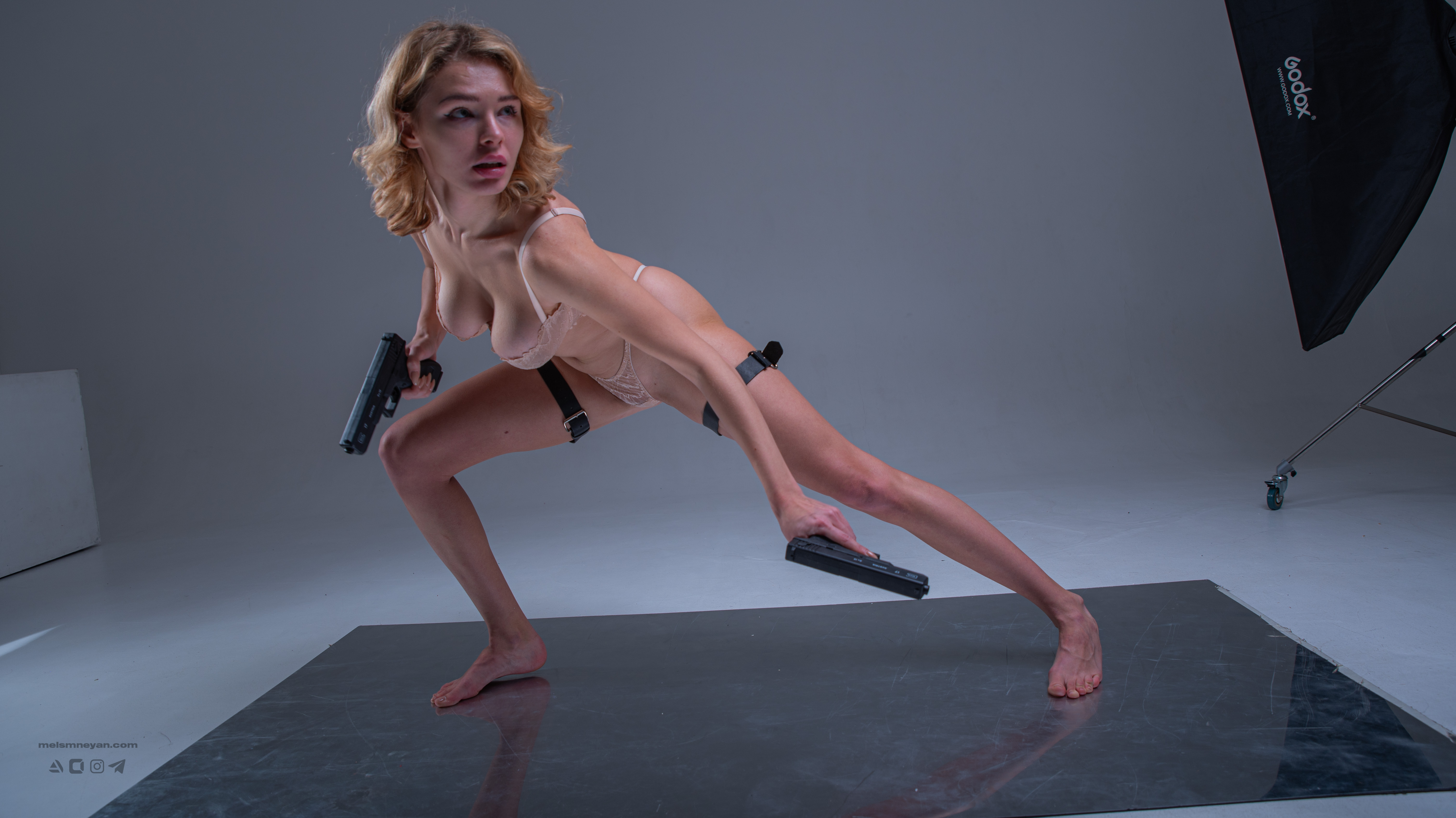a woman in lingerie poses with two guns in a studio setting. the woman is barefoot and wearing straps on her thighs. she is holding a gun in each hand and has a focused expression on her face. a softbox with "godox" logo is visible in the background.