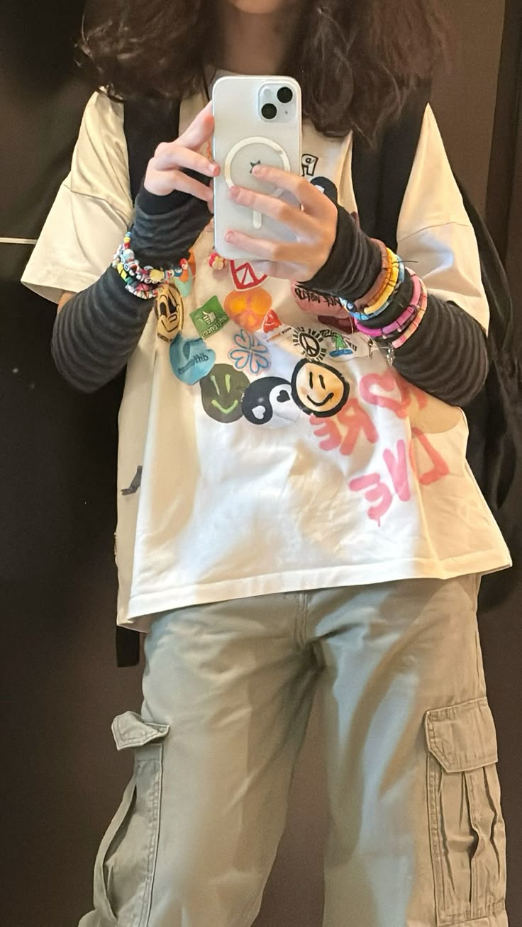 a person is taking a selfie. the person is a young female with long, curly hair. she is wearing a white tshirt with colorful graphics and the words "make love" on it, with long striped sleeves underneath. she has several colorful wristbands and bracelets on her wrists, and a necklace with pins. she is also wearing baggy cargo pants. she is holding an iphone in her left hand, and a backpack is slung over her shoulder. the background is a plain wall.