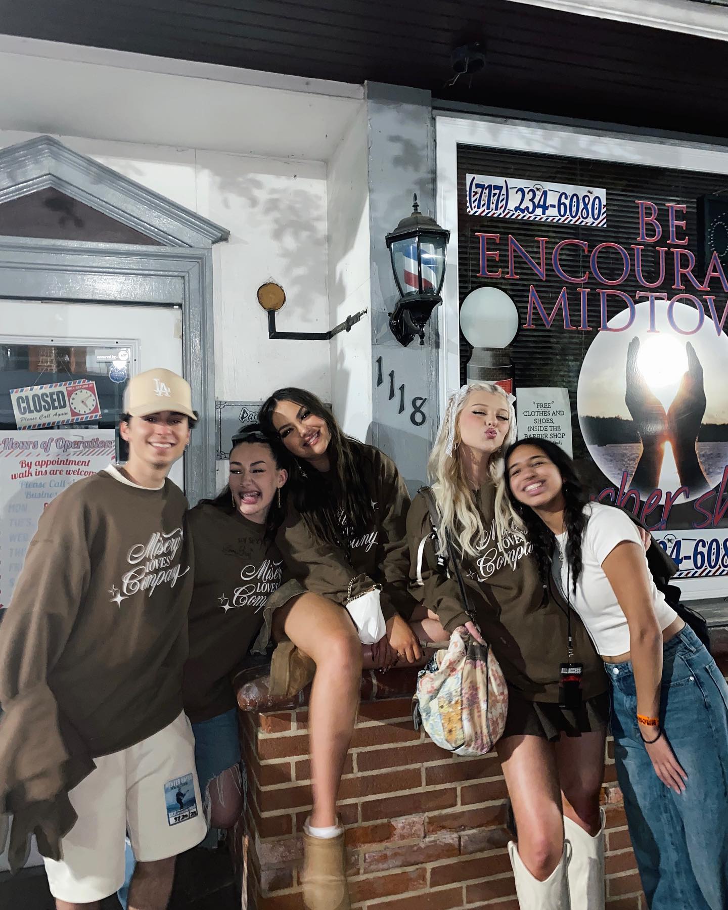 a group of five young people pose outside a building with a barbershop sign. they are standing and sitting on a low brick wall. the person on the left wears a brown sweatshirt, white shorts, and a baseball cap. the next two wear similar brown sweatshirts, one paired with jeans and the other with ugg boots. the next person wears a similar sweatshirt with a short skirt and white boots, holding a patterned bag. the person on the right is wearing a white tshirt and jeans. the background includes a storefront with a "closed" sign and a barbershop sign that reads "be encouraged midtown".