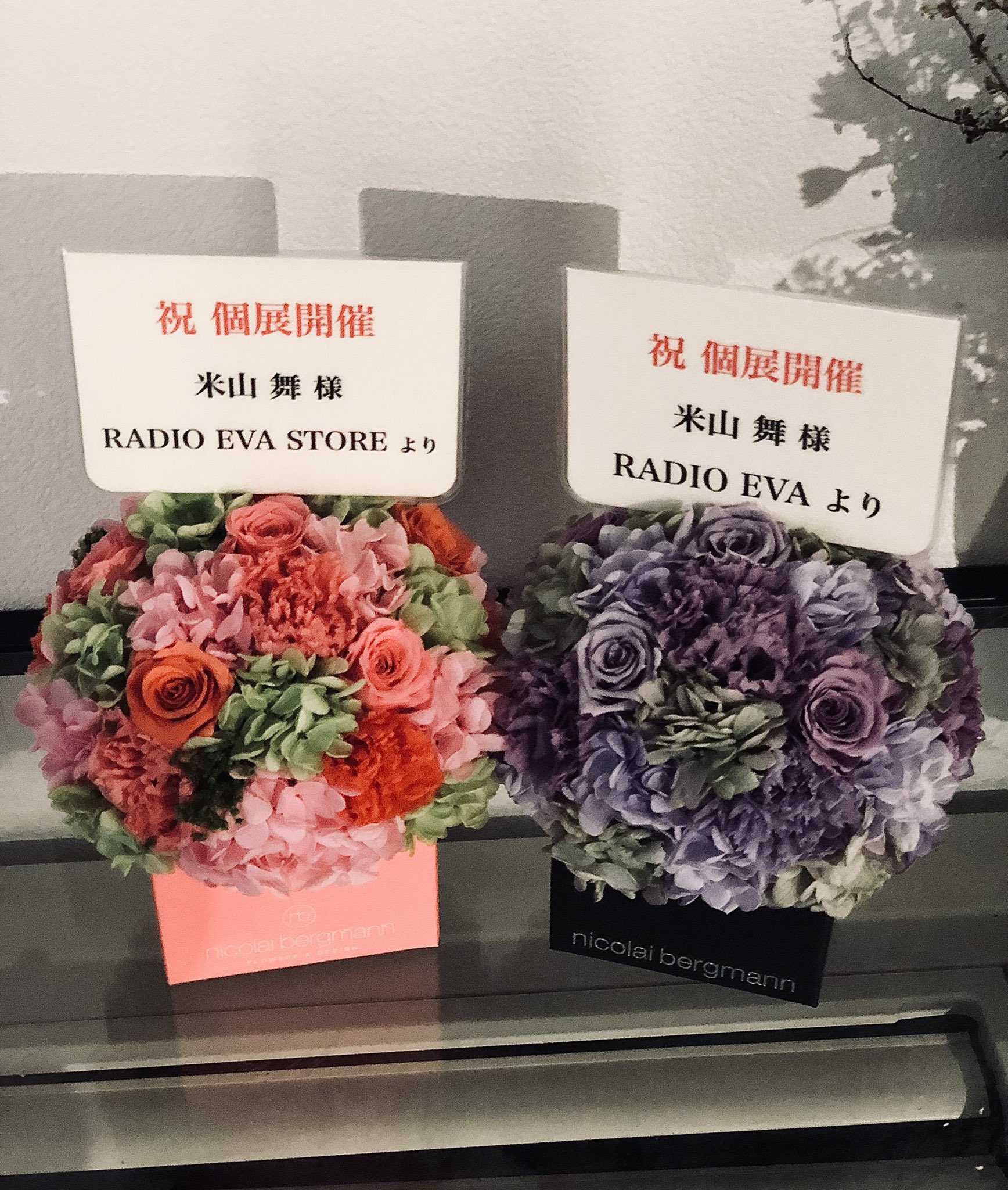 wo bouquets of flowers, one pink and one purple, are displayed on a counter. each bouquet is accompanied by a card congratulating someone named 米山舞 on their solo exhibition.  the cards indicate the bouquets are gifts from radio eva and radio eva store. the purple bouquet is in a box labeled "nicolai bergmann".
