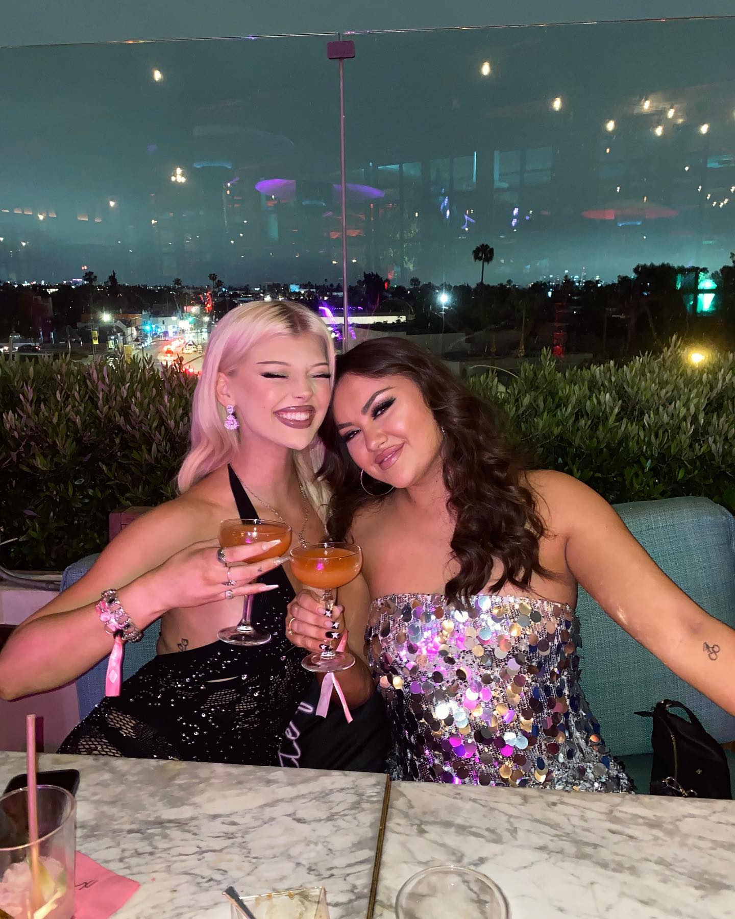 two women are posing for a photo while holding drinks at a rooftop bar at night. the woman on the left has blonde hair and is wearing a black dress. the woman on the right has dark