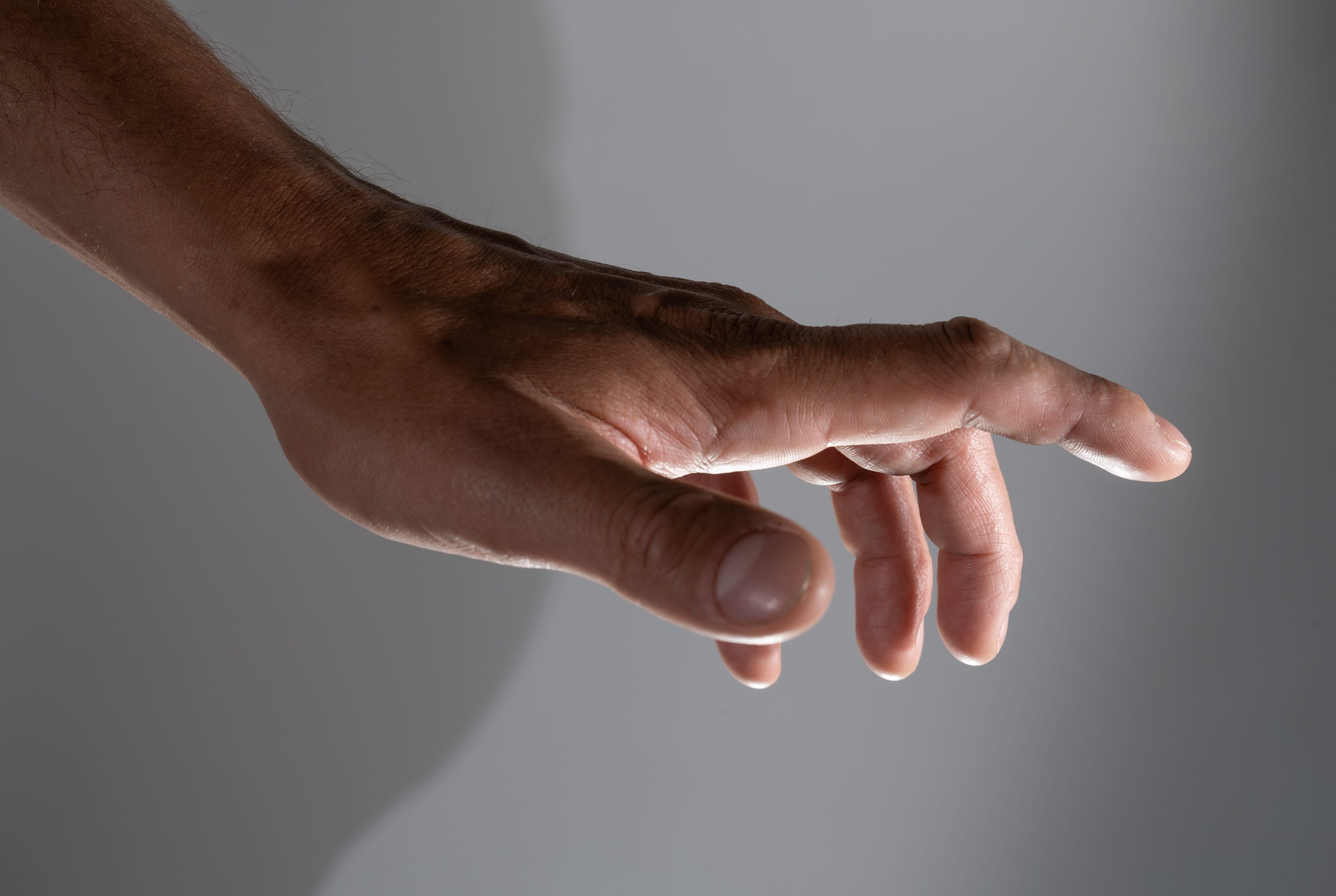 a closeup side view of a man's outstretched hand against a neutral grey background. the hand is reaching forward, fingers slightly curled. the lighting is soft and natural, coming from the side, which creates subtle shadows and highlights the texture of the skin.