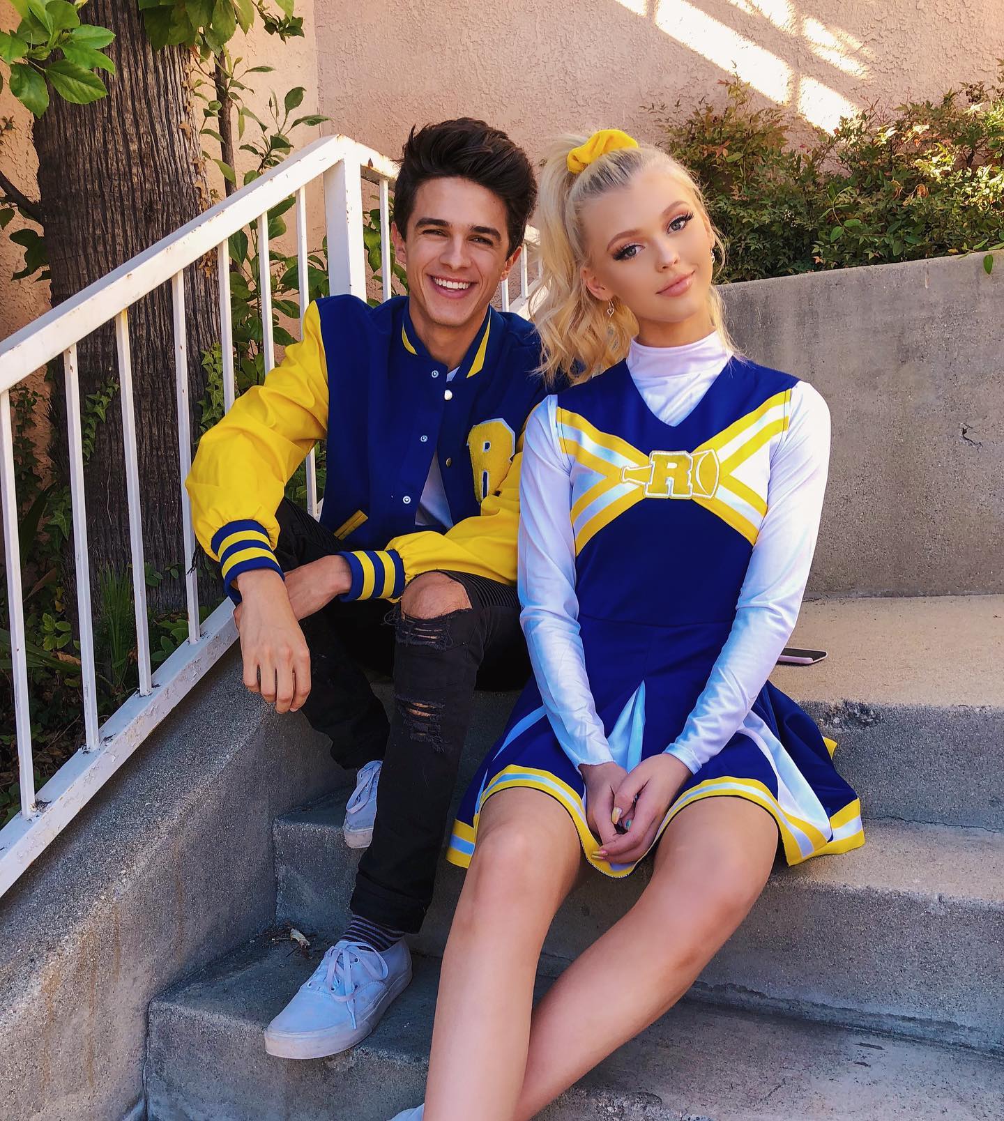 a young man and woman are sitting on outdoor concrete stairs. the man is wearing a blue and yellow letterman jacket, ripped jeans, and light blue sneakers. the woman is wearing a blue and yellow cheerleading uniform, with a high blonde ponytail. both are smiling and look to be enjoying themselves. there are trees and plants in the background.