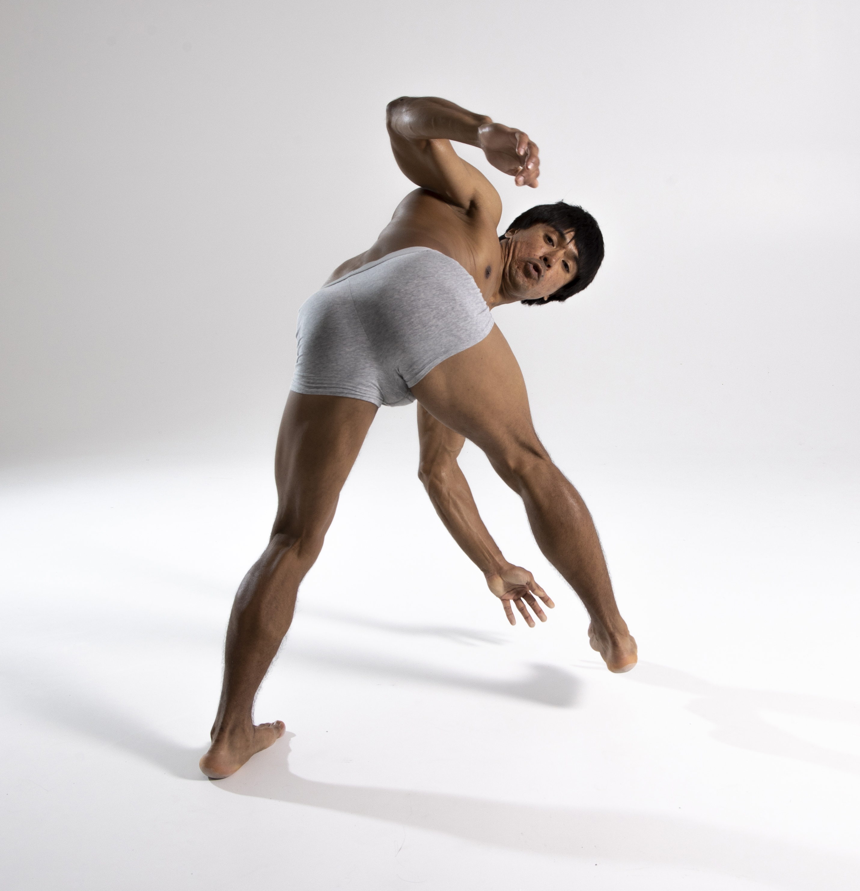 a muscular, barechested man in gray underwear is captured in a dynamic, athletic pose against a bright white background. he appears to be a dancer or athlete demonstrating flexibility and movement.