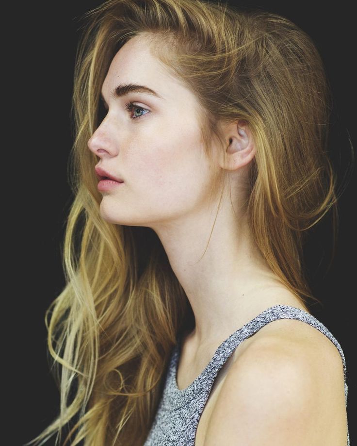 a profile portrait of a woman with blonde hair, blue eyes, and fair skin wearing a gray tank top against a black background.