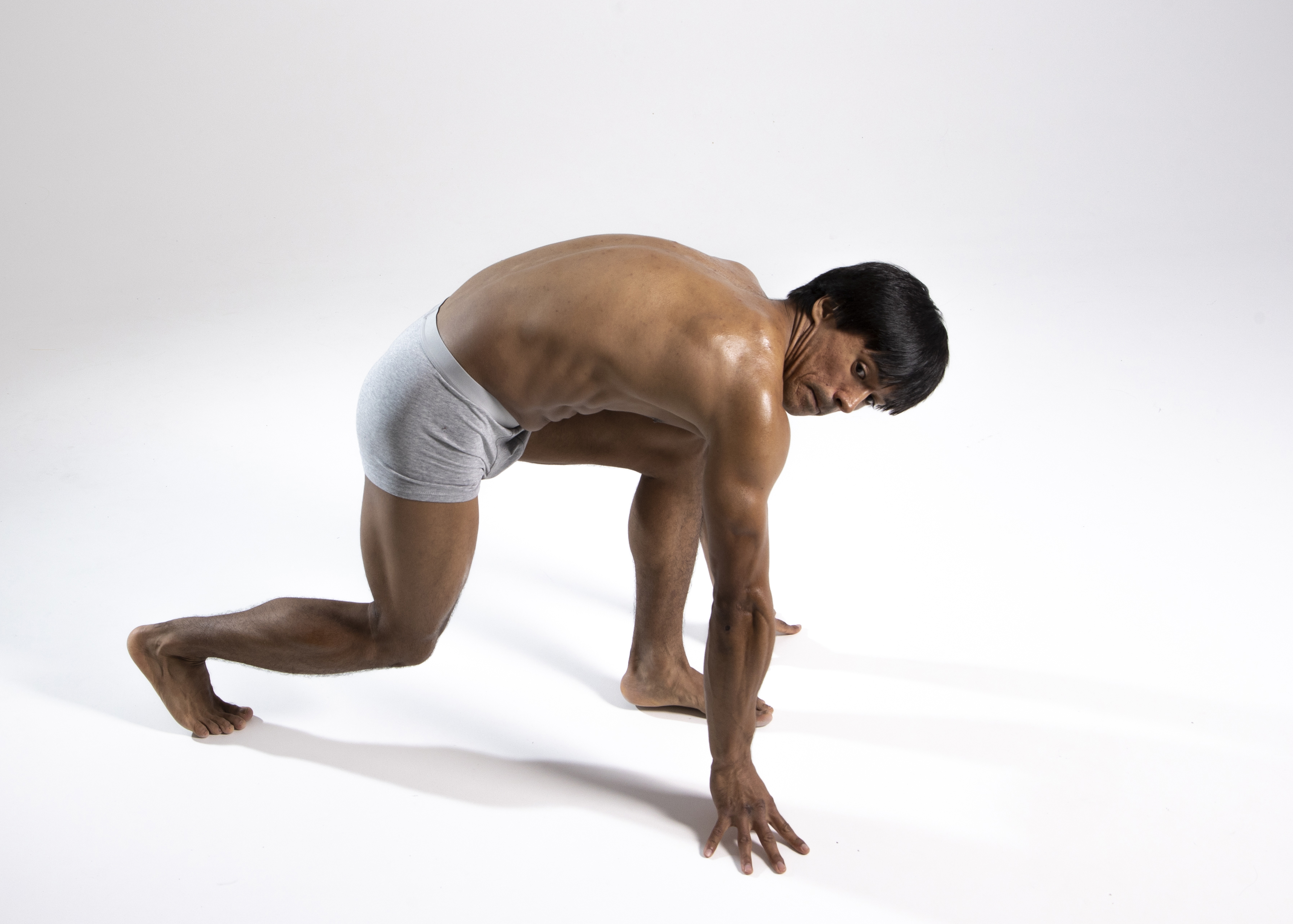 an athletic man is in a starting position on a white background. he is wearing gray underwear and is barefoot. his skin appears tanned and he has black hair. he is crouching with his hands on the ground, looking down. his body is muscular. he is isolated in a studio shot.