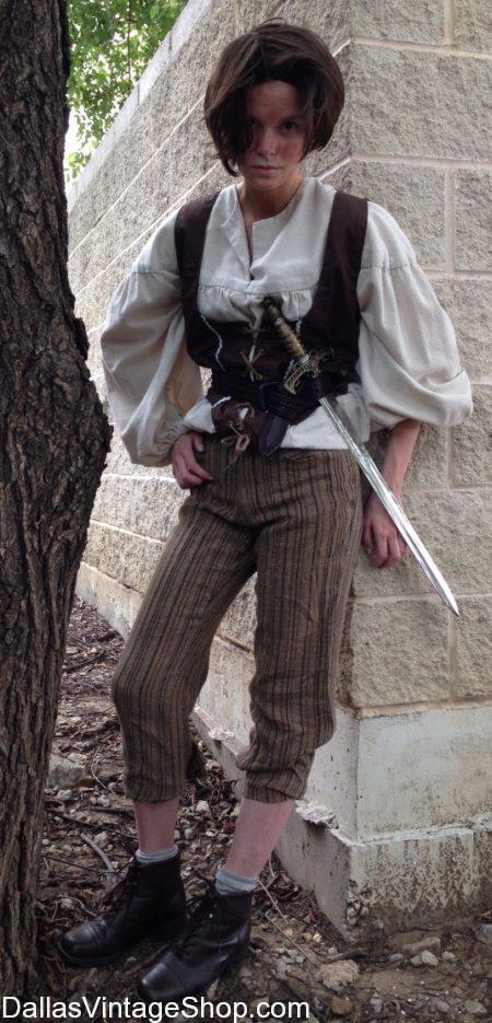 a woman dressed in a historical costume stands in front of a wall. she is wearing a white shirt, brown pants, and black boots. she is holding a sword in her right hand. she is looking at the camera. the image is taken outdoors during the day.