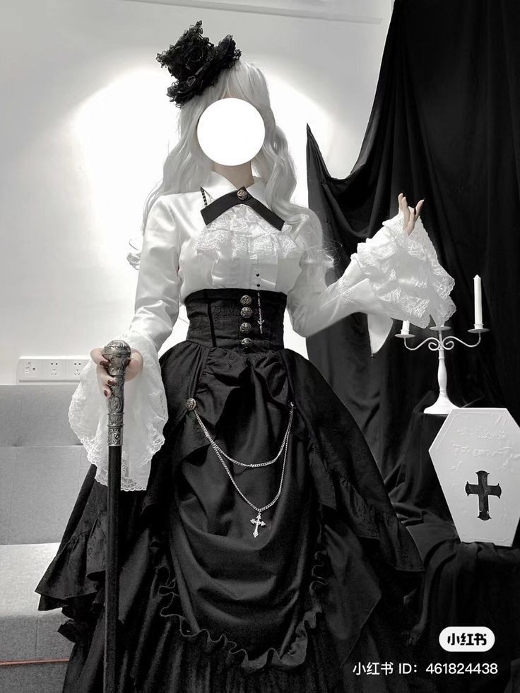 a woman wearing a black and white gothic victorian dress stands holding a walking stick. the dress has a fitted black corset and a long, ruffled skirt. she wears a white blouse with lace detailing and large, ruffled sleeves. a silver cross necklace hangs from her waist. she has a white wig and a small black hat. next to her is a small white coffin prop with a black cross on it and a candlestick with two candles. a black curtain hangs in the background.