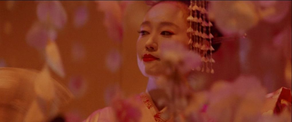 a woman dressed in a traditional japanese kimono stands amidst a background of blurred flowers and lights. she is looking to the side, her face partially hidden by a delicate floral pattern. her makeup is flawless, and her red lips are a striking contrast to her pale skin. the overall effect is one of elegance and mystery, creating a sense of beauty and intrigue.