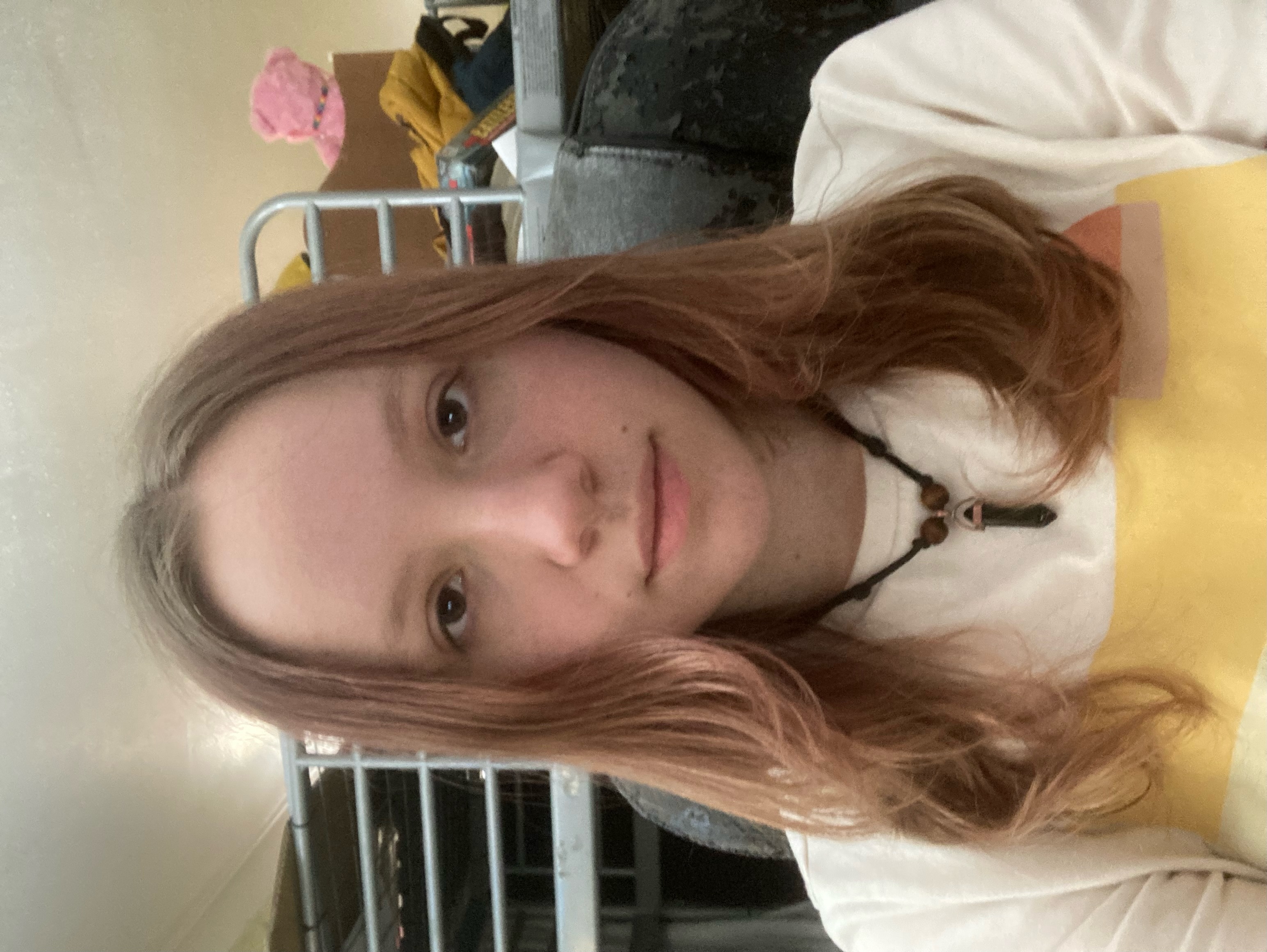 a young woman with long brown hair is taking a selfie. she is wearing a white tshirt with a yellow design and a necklace with a black crystal pendant. she has a neutral expression on her face. behind her, a bunk bed is visible.  the photo is taken with natural light coming from the side.