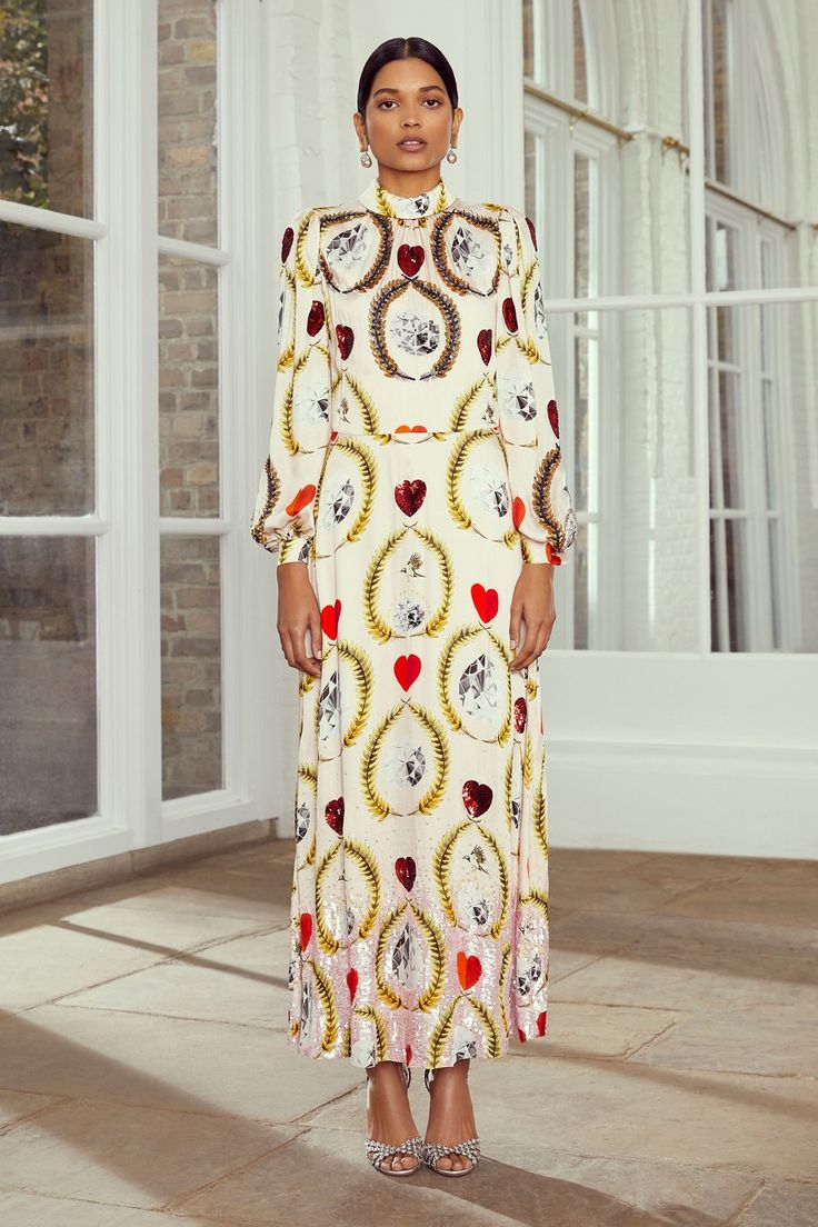 a fulllength shot of a woman standing in a long, white maxi dress. the dress features a high neck, long sleeves, and a repeating pattern of hearts, floral elements, and small pictures within decorative frames. the dress also appears to incorporate sequins adding a bit of shimmer to the garment. the dress is elegant and stylish. the woman is wearing highheeled sandals and pearl earrings, adding to her sophisticated appearance. she is standing on a lightcolored floor in what appears to be a bright interior space. the setting seems elegant and sophisticated, fitting the style of the clothing.