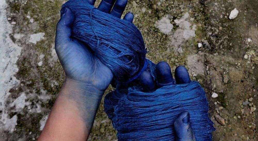 a pair of hands, stained blue from indigo dye, hold a thick bundle of deep blue yarn. the background is a rough, textured ground with hints of blue dye splattered on it. the image suggests the process of handdyeing yarn with natural indigo.