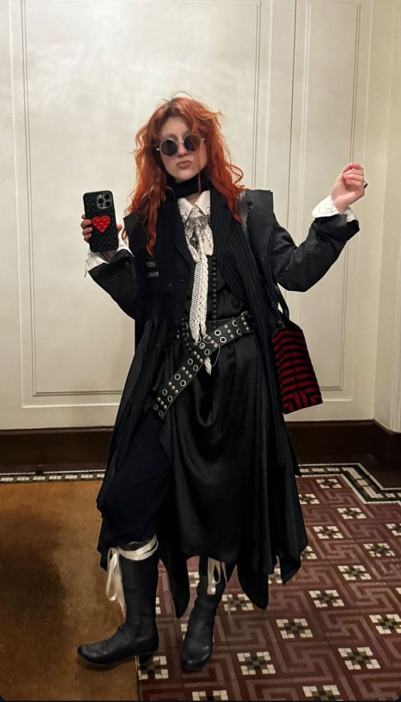 a young woman with long red hair and a black coat stands on a tiled floor.  she is wearing sunglasses, black boots, a black dress, a white shirt, and a black bag.  she is posing for a photograph.