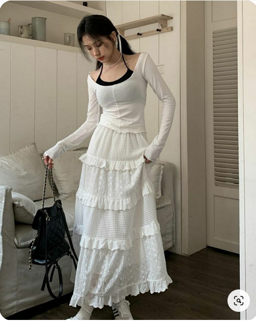 a young woman is posing for a mirror selfie indoors. she is wearing a white twopiece outfit that features a longsleeved, squareneck top and a tiered midi skirt with ruffles. she is also wearing white sneakers and carrying a black shoulder bag. the lighting is bright and natural, and the perspective is eyelevel.