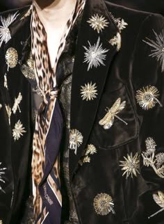 a closeup shot of a man's blazer with intricate floral embroidery and a leopard print scarf. the blazer is black velvet, with a subtle sheen, and the embroidery is in gold and silver thread. the scarf is tied loosely around the neck, with the ends tucked inside the blazer. the overall look is elegant and refined, with a touch of vintage flair. the image captures the details of the garment, highlighting the craftsmanship and beauty of the design.