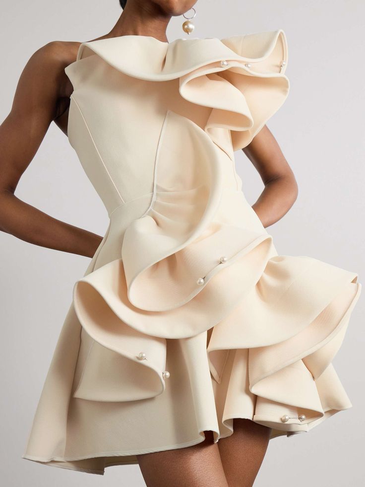a woman in a white dress, with pearl embellishments on the ruffles, poses in a studio. the dress is oneshoulder and features a dramatic ruffle design. the woman's hair is not visible, and she is wearing pearl earrings. the overall look is elegant and formal.