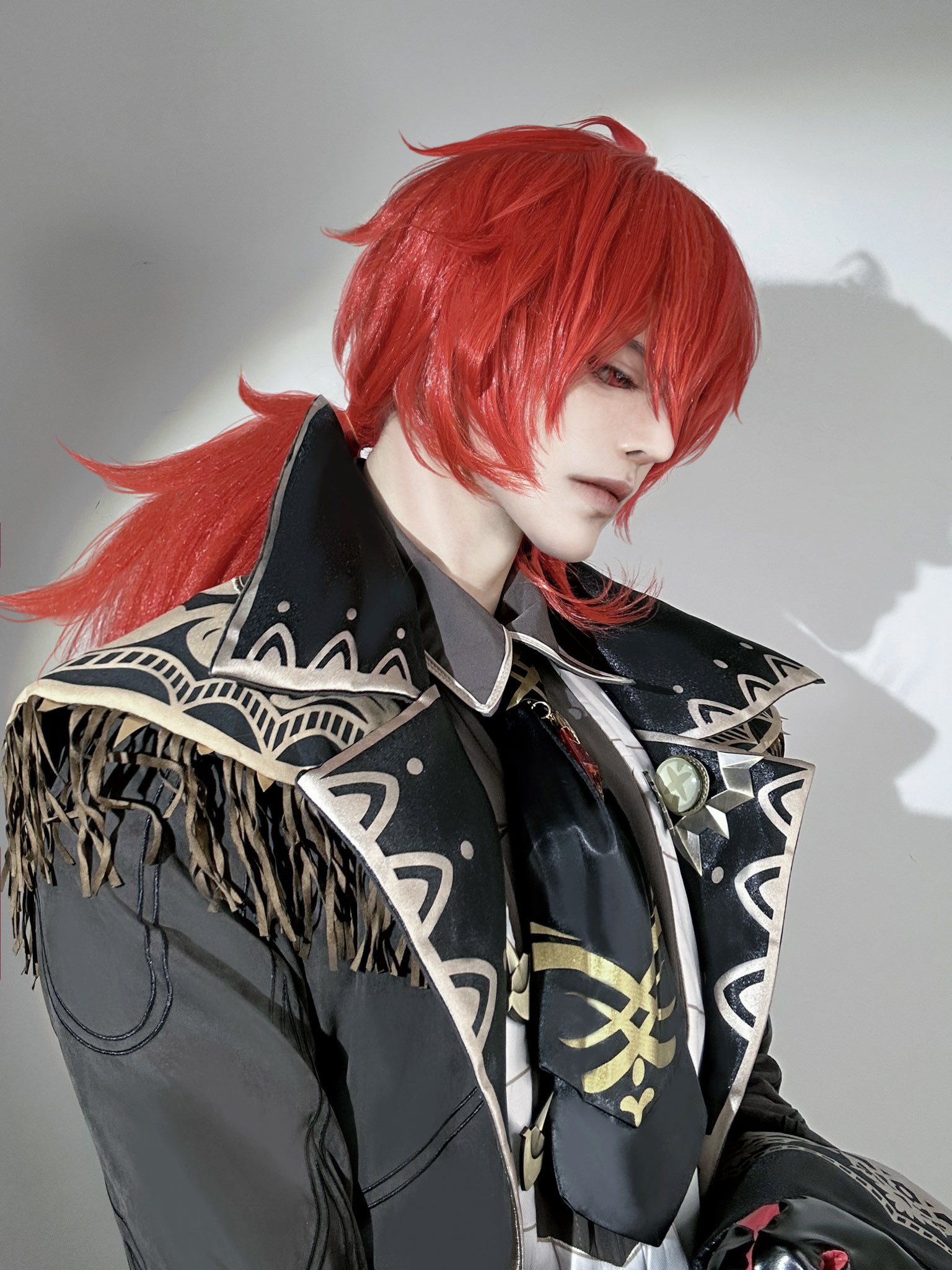 a man with red hair in a cosplay costume, wearing a black jacket with gold accents,  and a black tie. the background is white and he is looking down.  he is portraying a male character from an anime show.