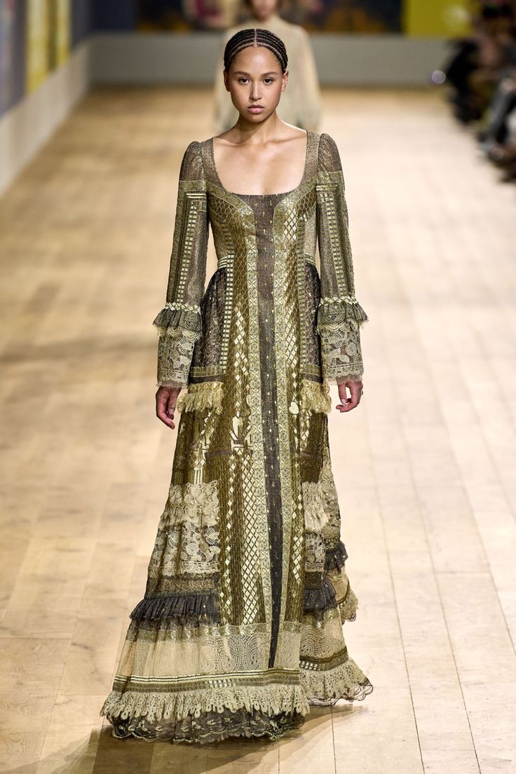 a full shot of a darkskinned model walking down a runway wearing a long, elaborate gown.  the dress is a muted olive green and gold, featuring intricate embroidery, lace detailing, and layers of ruffles. the model's hair is styled in a neat updo. the background is blurred, showing a wooden floor and a glimpse of other models in the distance. the overall aesthetic is high fashion, with a bohemian or vintage feel.