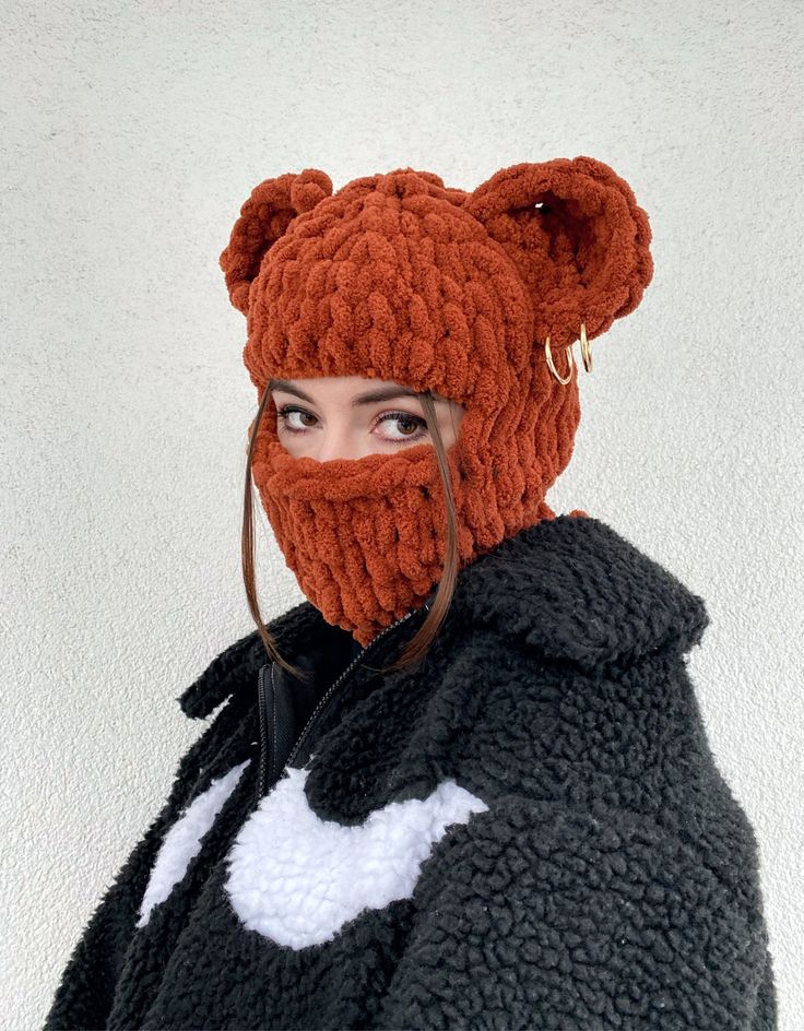 a woman is wearing a brown knit hat with bear ears that covers her face. the hat is warm and fuzzy and looks very comfortable. the woman is wearing a black jacket with white fur trim. the background is a plain white wall. she looks stylish and ready for winter.