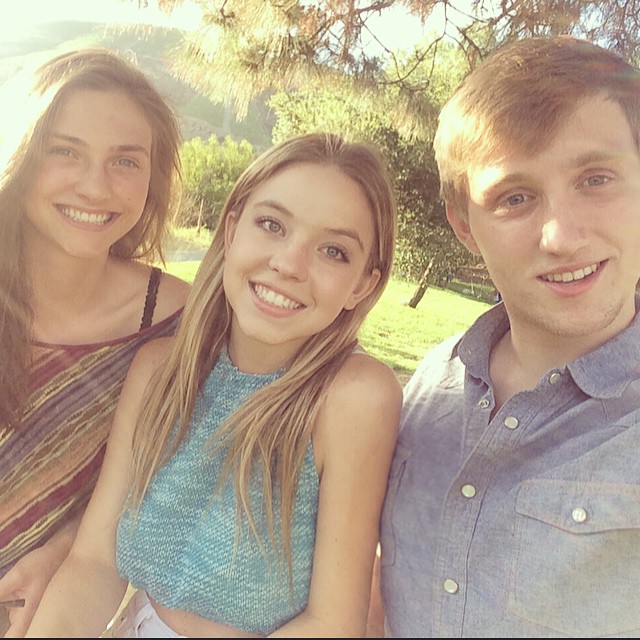 the image shows three young adults, two girls and a boy, posing closely together outdoors in what appears to be a park.  the girls are smiling brightly at the camera. one girl has long blonde hair and is wearing a teal knitted top, the other girl has long brown hair and is wearing a multicolored striped shirt.  the boy has light brown hair and is wearing a light blue buttondown shirt. they are standing in a sunny area with trees and greenery in the background. the overall mood is cheerful and suggests a friendly gathering.