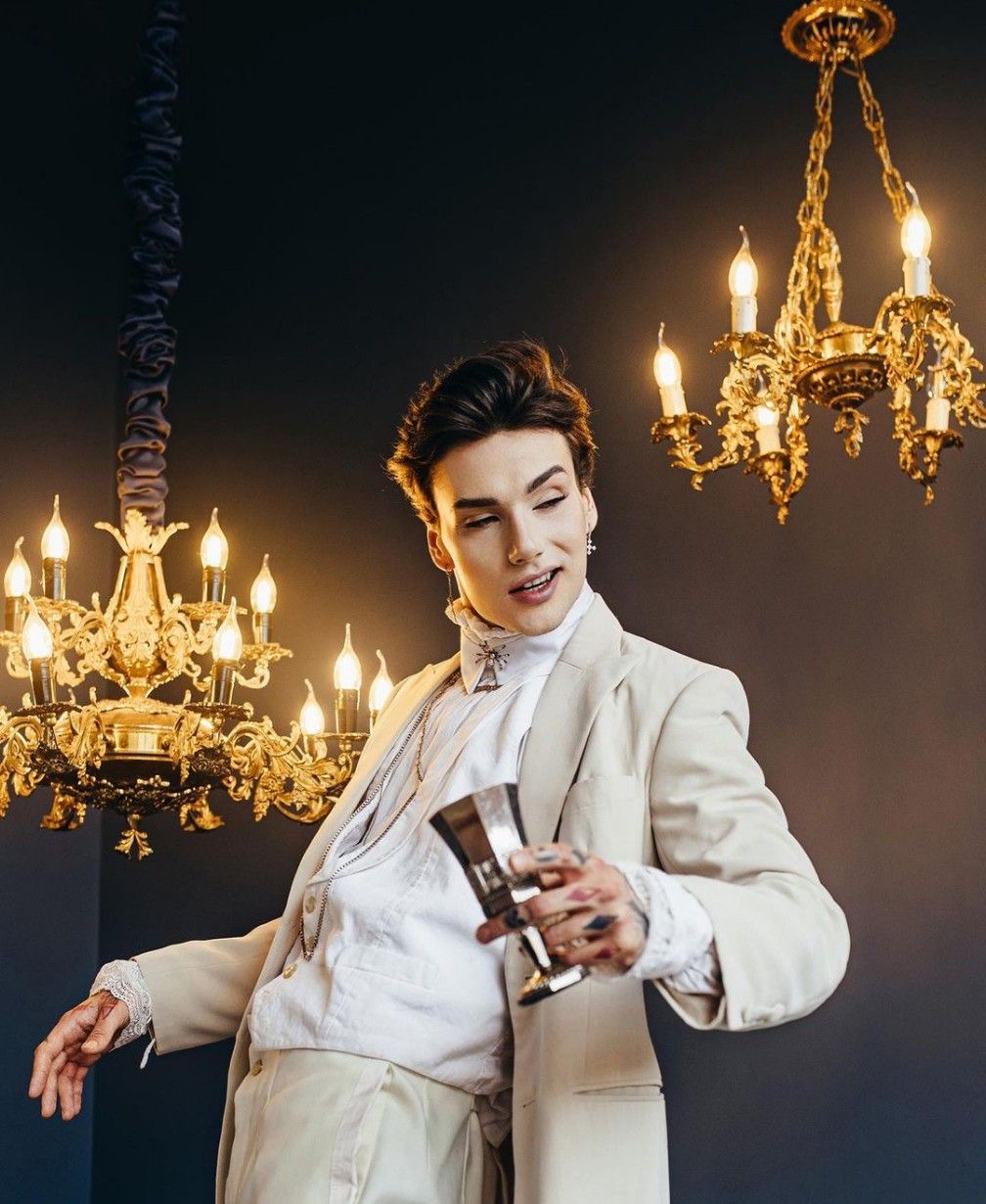 a man in victorianstyle clothing is standing in front of a dark background. he is holding a goblet and looking down at it. there are two ornate chandeliers on either side of him, illuminating the scene with warm light.  his outfit has a whimsical air,  evoking a gothic fantasy aesthetic.