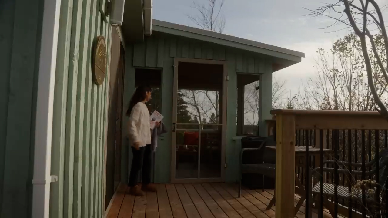 a woman stands on a wooden deck outside a light blue cabin. the deck leads to a screenedin porch with furniture visible inside.  the cabin is surrounded by trees with colorful fall foliage. the scene is bathed in warm, natural sunlight.