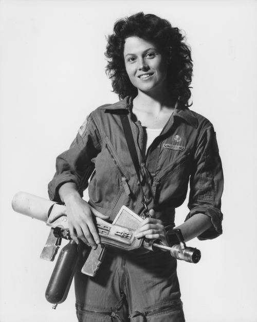 this is a black and white portrait of a woman. she is looking at the camera and smiling. the woman is dressed in a grey jumpsuit and is holding a futuristic looking gun.  she looks like a character from a science fiction film.