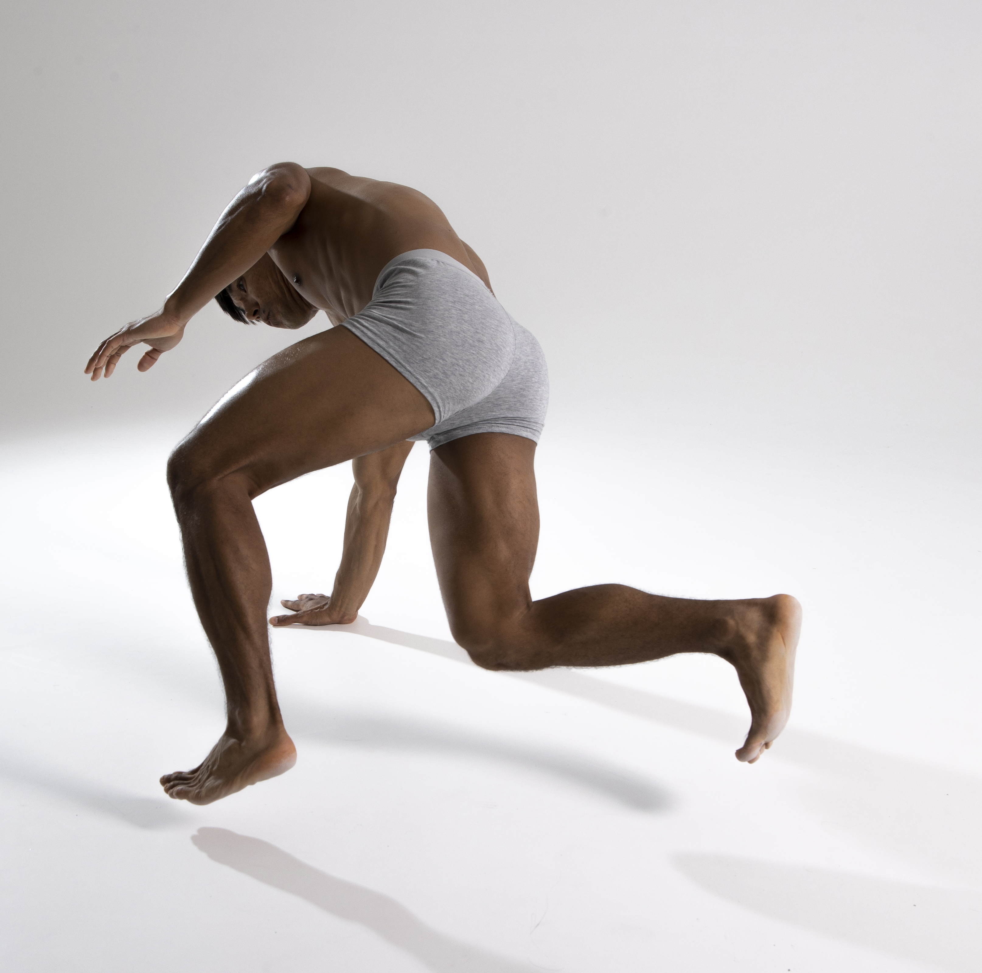 a darkskinned, muscular man wearing grey underwear is crawling on a white surface in a studio setting. he is barefoot and the pose emphasizes his physique.