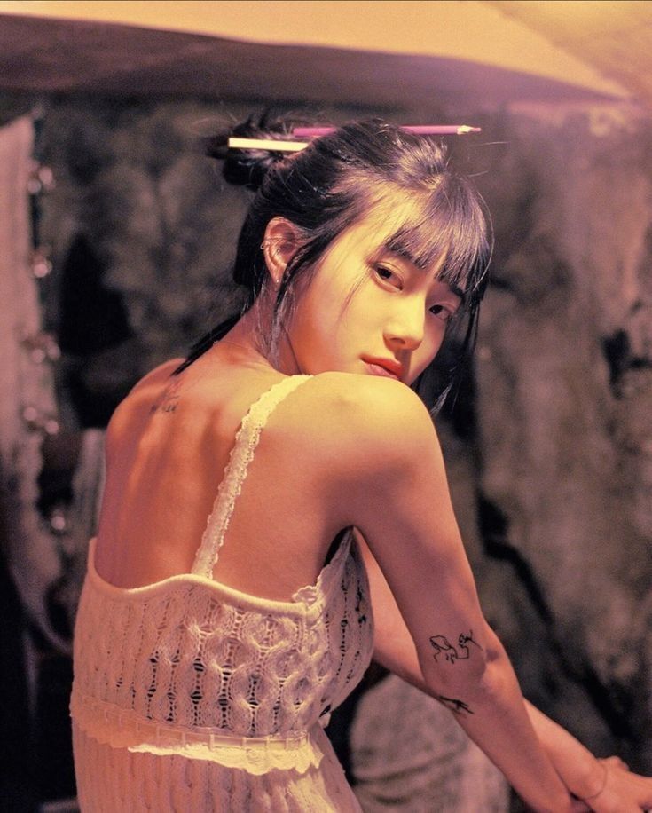 an asian woman with bangs and her hair in an updo with a pencil through it is looking over her shoulder at the camera. she is wearing a white tank top, and there is a tattoo on her arm and back. the background is out of focus.