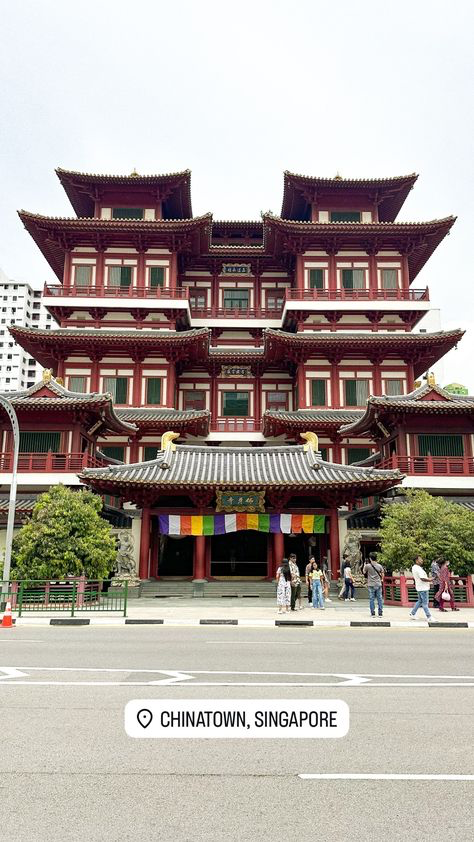 the image shows the buddha tooth relic temple and museum in chinatown, singapore.  it's a multistory red building with traditional chinese architecture, featuring intricate details and layered roofs.  the street in front of the temple is relatively quiet with a few people walking around. the image is taken from across the street, providing a full view of the temple's facade. a small location marker indicating "chinatown, singapore" is present at the bottom of the image.