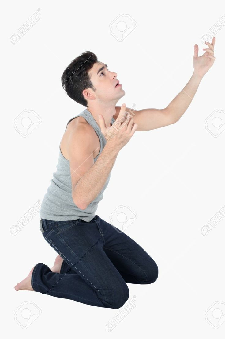 a young caucasian man is kneeling on the floor, looking up at the ceiling with a desperate and hopeful expression on his face. he is wearing a grey tank top and jeans and his arms are raised in a pleading gesture. the background is a plain white wall.
