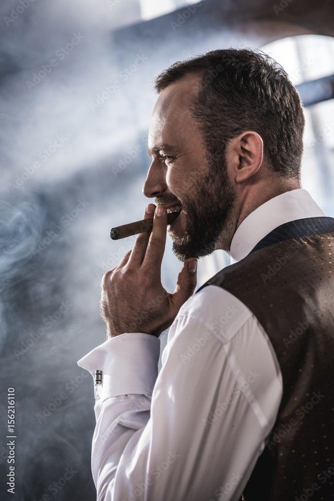 a close up, portrait of a handsome, bearded man, wearing a white shirt and suit vest. the man is holding a cigar and smoking. he is looking away from the camera and appears pensive, contemplative and relaxed.  the portrait is moody and dark, shot in a studio setting.
