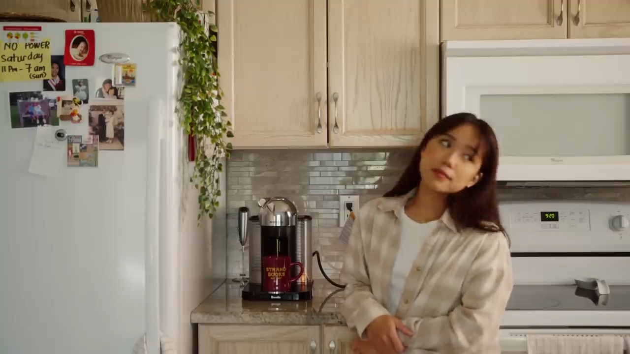 a young woman stands in a cozy kitchen with a soft, warm color palette.  she is casually leaning against the counter, looking to the side. a coffee maker, a mug, and a leafy green plant are visible on the counter behind her. the kitchen features light wood cabinets, a white refrigerator covered in photos and notes, and a white microwave. the overall atmosphere suggests a relaxed, domestic scene, possibly early in the morning.