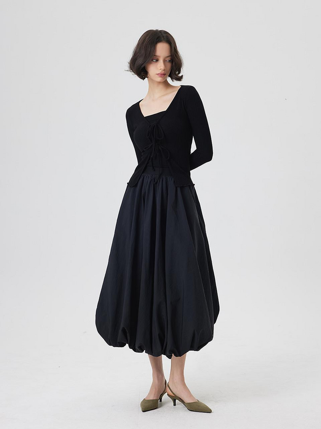a woman stands in a studio wearing a black top with long sleeves and a square neckline with tie details. she is wearing a puffy black midi skirt and olive green heels. the background is white.