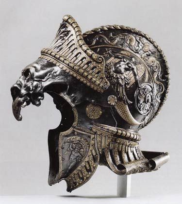 this is a closeup view of an ornate, antique helmet, crafted from a dark metal. the helmet, likely dating back to the medieval or renaissance period, features intricate engravings and embellishments. the design showcases a fierce, grotesque face with a gaping maw, possibly intended to intimidate during battle.  this piece appears to be a museum artifact, carefully lit to highlight its craftsmanship and details against a neutral background.