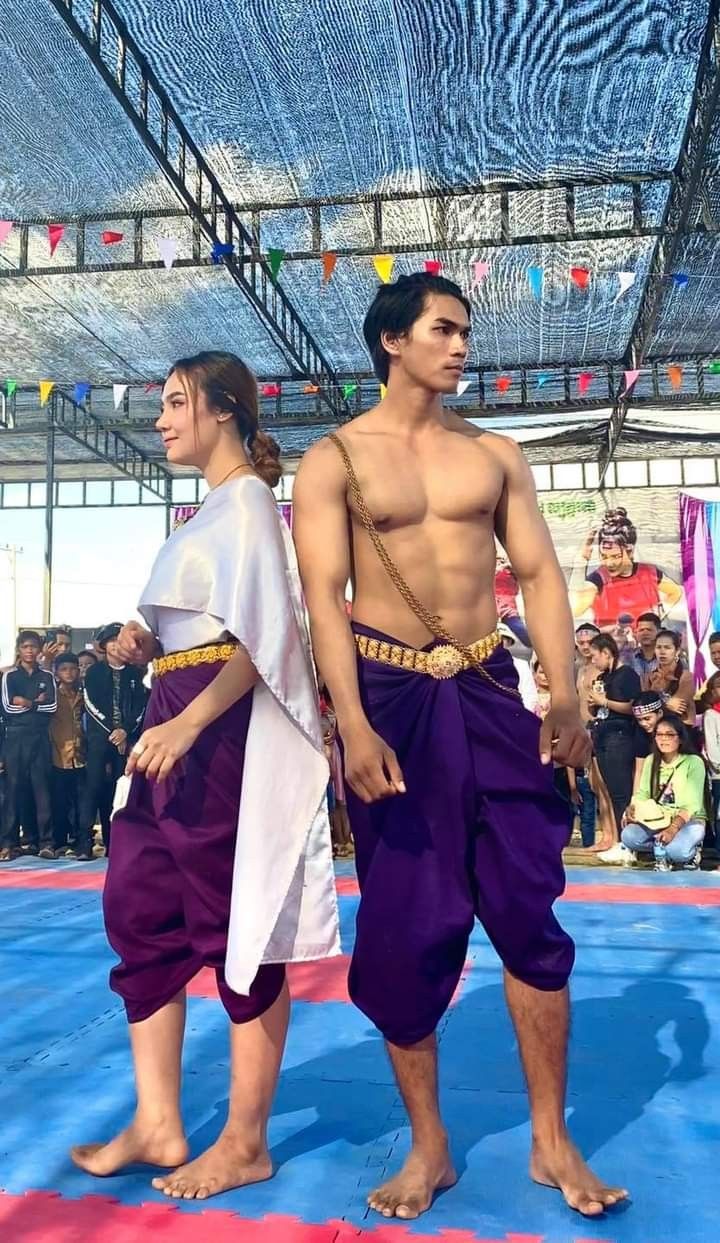 a full shot of a man and a woman dressed in traditional cambodian attire stand on a blue mat outdoors.  the man, shirtless, wears a purple sampot (loincloth) and a gold waist sash. the woman wears a purple sampot and a flowing white top. both are barefoot.  they appear to be at an event or performance, with spectators visible in the background under a shaded area. colorful flags hang from above. the man is muscular and has an athletic build. the scene is brightly lit.