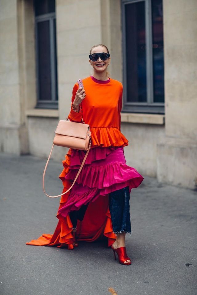 a woman is walking on a street, wearing a bright orange and pink ruffled dress over dark denim jeans, paired with red heeled mules. she carries a light pink handbag over her shoulder and holds a smartphone in her hand. she's also wearing black sunglasses and smiling. the background shows a building with windows and a neutralcolored facade.