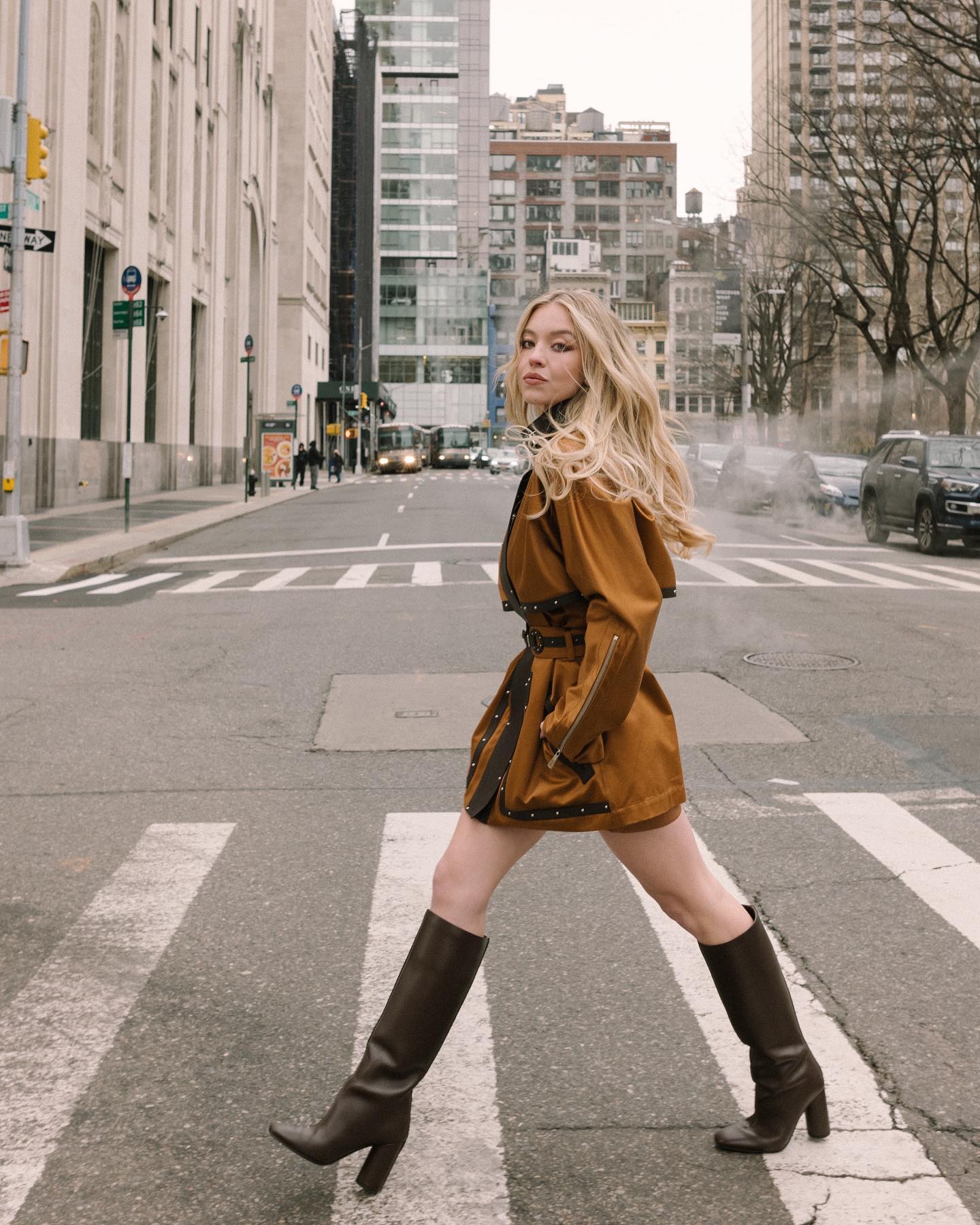 a stylish blonde woman in a brown trench coat and brown high boots walks across a city street in new york city.  she's crossing a pedestrian crossing, and the city buildings form a backdrop. the overall style is fashionable and urban. the image is taken during the day in natural light.