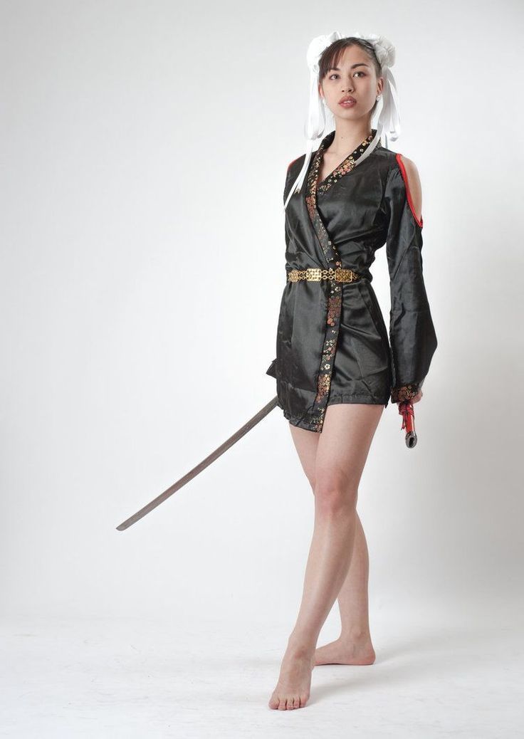 a young woman is standing in a white studio, looking at the camera. she is wearing a traditional japanese kimono with a black and gold pattern. she is also wearing a white headband and has a sword at her side. she is posing with a confident expression.