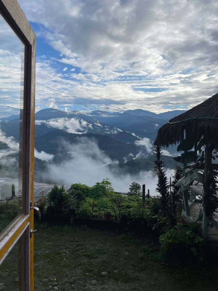 a scenic view of mountains with clouds enveloping the lower slopes, seen from an open window. a hut with a thatched roof is visible in the foreground, surrounded by lush greenery and vegetation. the sky is partly cloudy with patches of blue. the photo captures a natural, outdoor landscape.