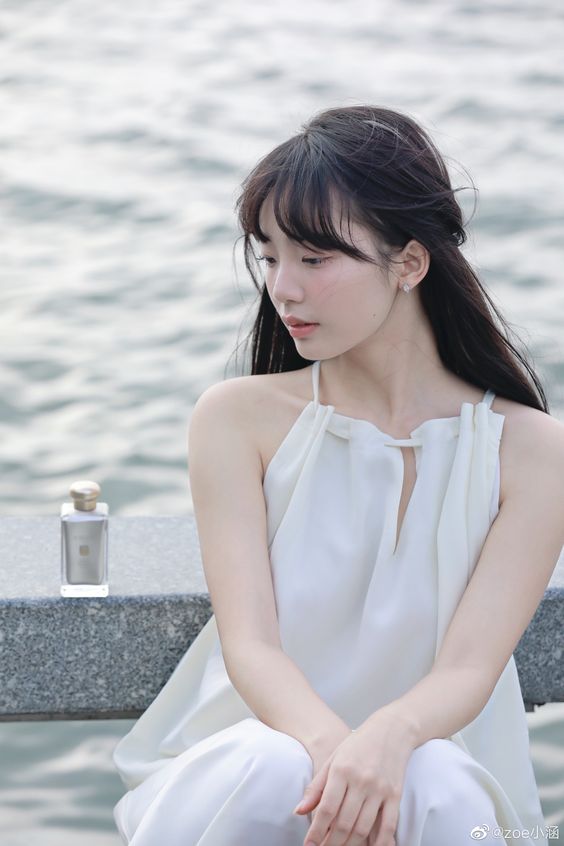 a young woman with black hair is sitting on a concrete ledge by a body of water. she is wearing a white halterneck dress and is looking down at a bottle of perfume beside her. she has a contemplative expression on her face. the lighting is natural and the perspective is at eye level.