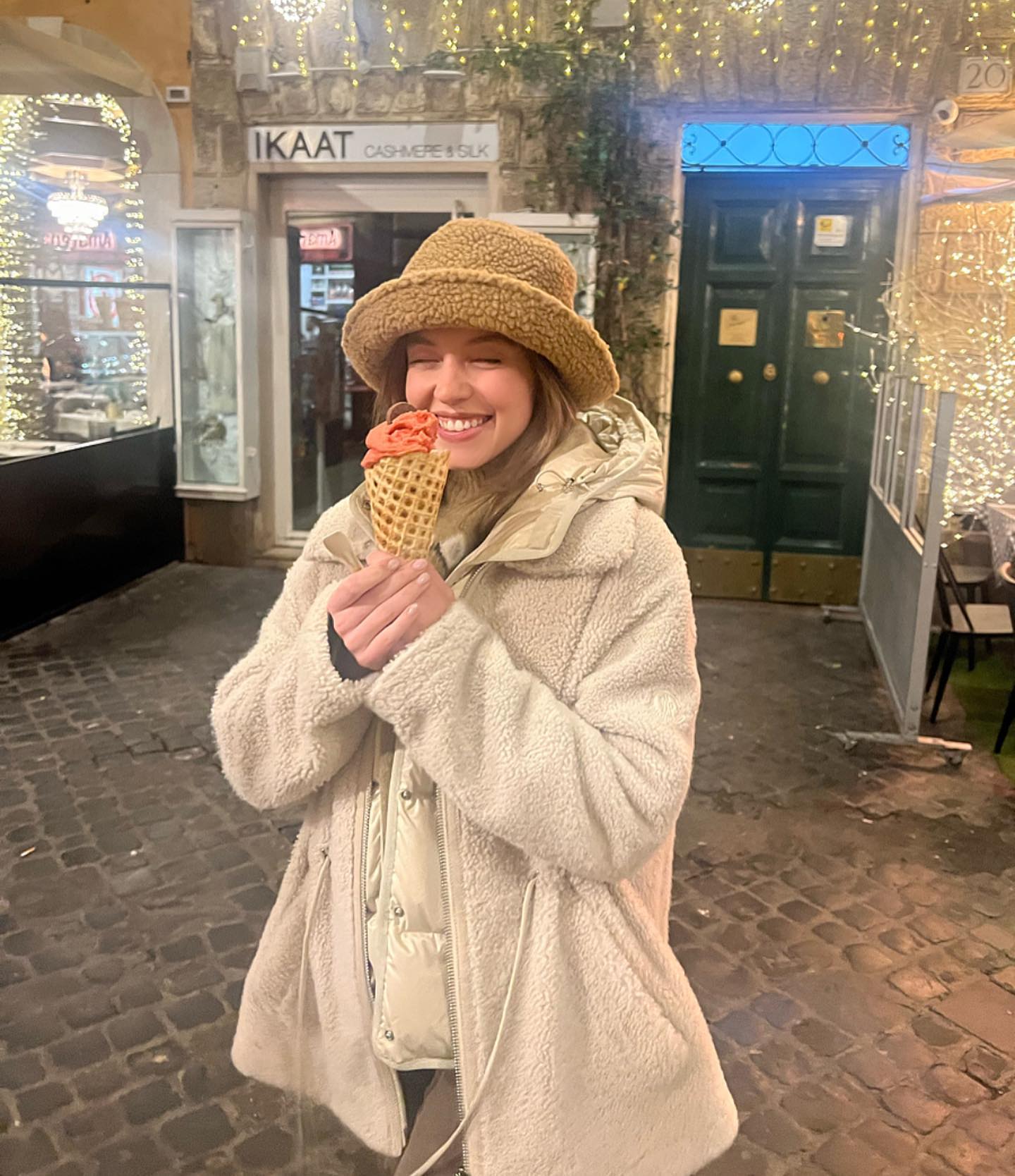 a young woman smiles happily while holding a double scoop of what appears to be strawberry gelato in a waffle cone. she is wearing a creamcolored teddy bear coat and a matching bucket hat. the setting is a charming european street at night with festive christmas lights in the background.  she seems to be enjoying her ice cream in a happy and relaxed way.