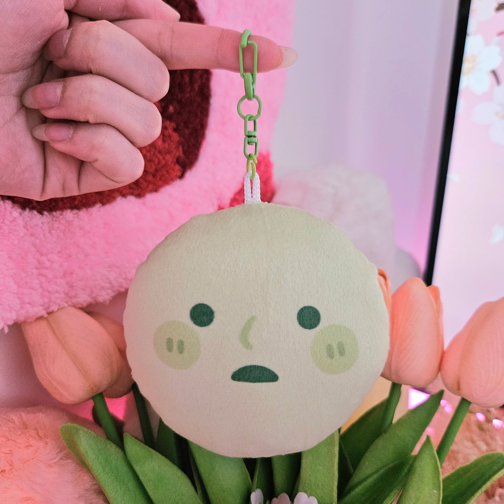 a closeup shot of a person's hand holding a small, round, pastelgreen plush toy keychain. the toy has a simple, cute face with green cheeks and a slightly sad expression. it's attached to a lightgreen chain. the background includes pink and peach tulips and a pink fuzzy blanket. the overall aesthetic is soft, pastel, and kawaii.