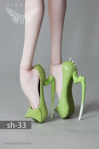 a closeup shot of a pair of green high heels on a doll's feet. the heels are a unique design, resembling a textured, possibly reptileinspired, appearance with a sharp, pointed texture. the background is a neutral gray, highlighting the vivid green of the shoes. the text "sh33" and "popovydolls.com" are visible in the lower part of the image. the image also features a butterfly logo in the top left corner.