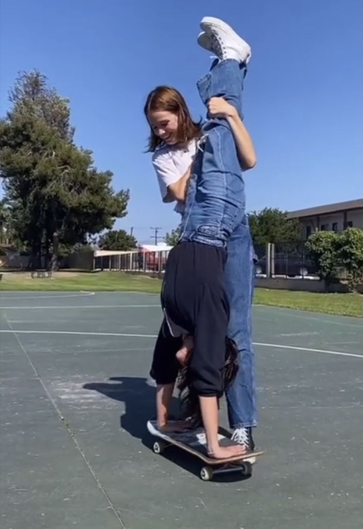 two girls are hanging out at the park and having fun. the girl in the front is doing a flip on a skateboard while the girl in the back is helping her. they are both smiling and enjoying their time together.