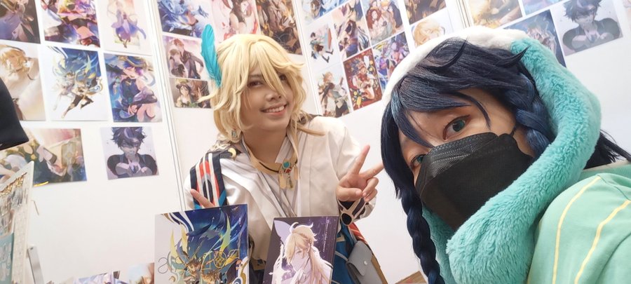 two cosplayers pose together in a friendly manner. one is cosplaying a character with blonde hair, a white and blue shirt, and a necklace. the other is cosplaying a character with blue hair, a light blue furry hat, and a face mask. both cosplayers seem very excited about the event and are smiling widely.  behind them, a wall covered with anime illustrations and posters can be seen.